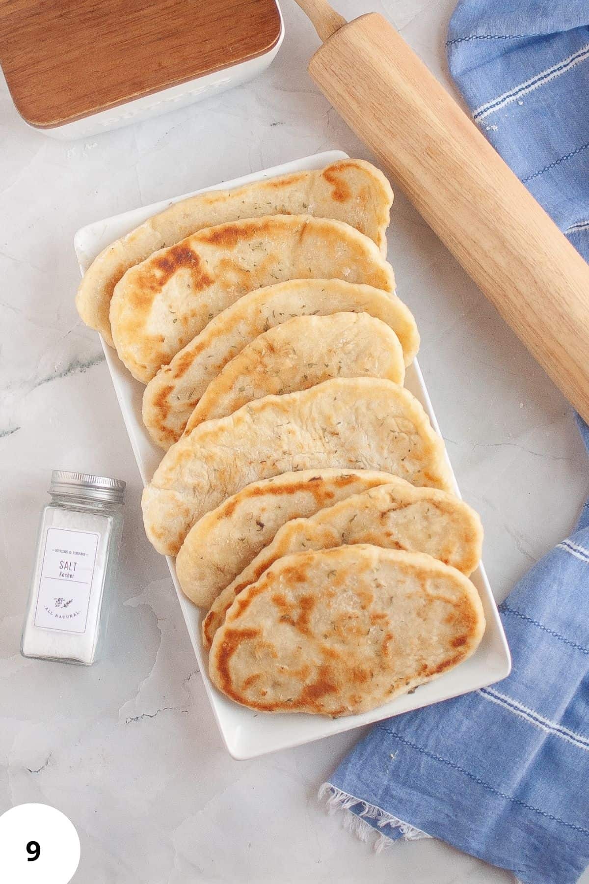 Freshly rolled naan neatly arranged in a serving dish with a rolling pin beside it on a tablecloth.