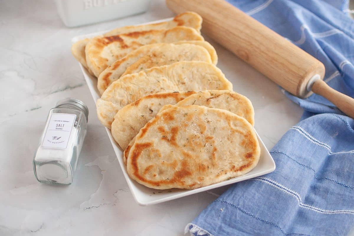 Freshly rolled naan neatly arranged in a serving dish with a rolling pin beside it on a tablecloth.