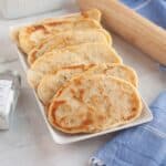 Freshly rolled naan neatly arranged in a serving dish with a rolling pin beside it on a tablecloth.