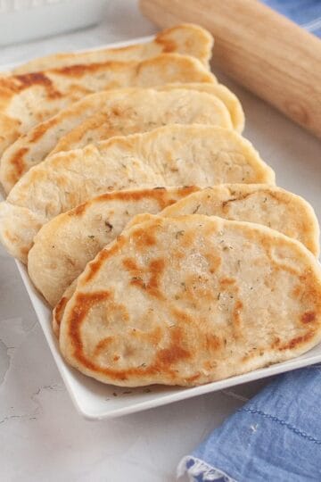 Freshly rolled naan neatly arranged in a serving dish with a rolling pin beside it on a tablecloth.