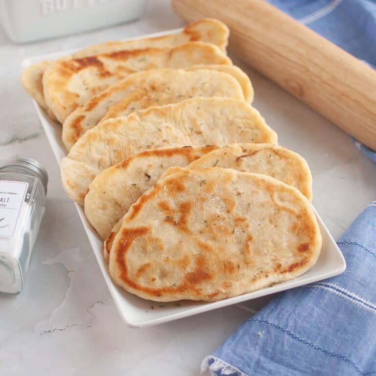 Freshly rolled naan neatly arranged in a serving dish with a rolling pin beside it on a tablecloth.