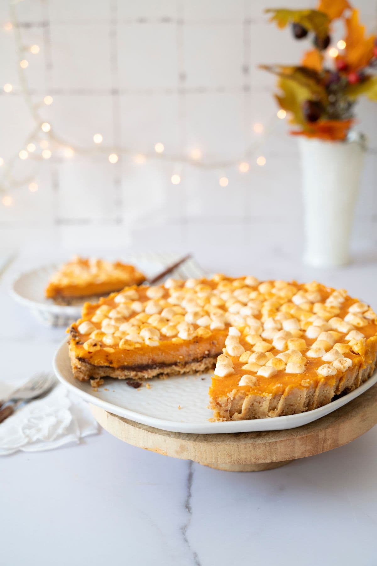 Whole sweet potato pie on a cake stand being sliced, with decorative lights in the background.
