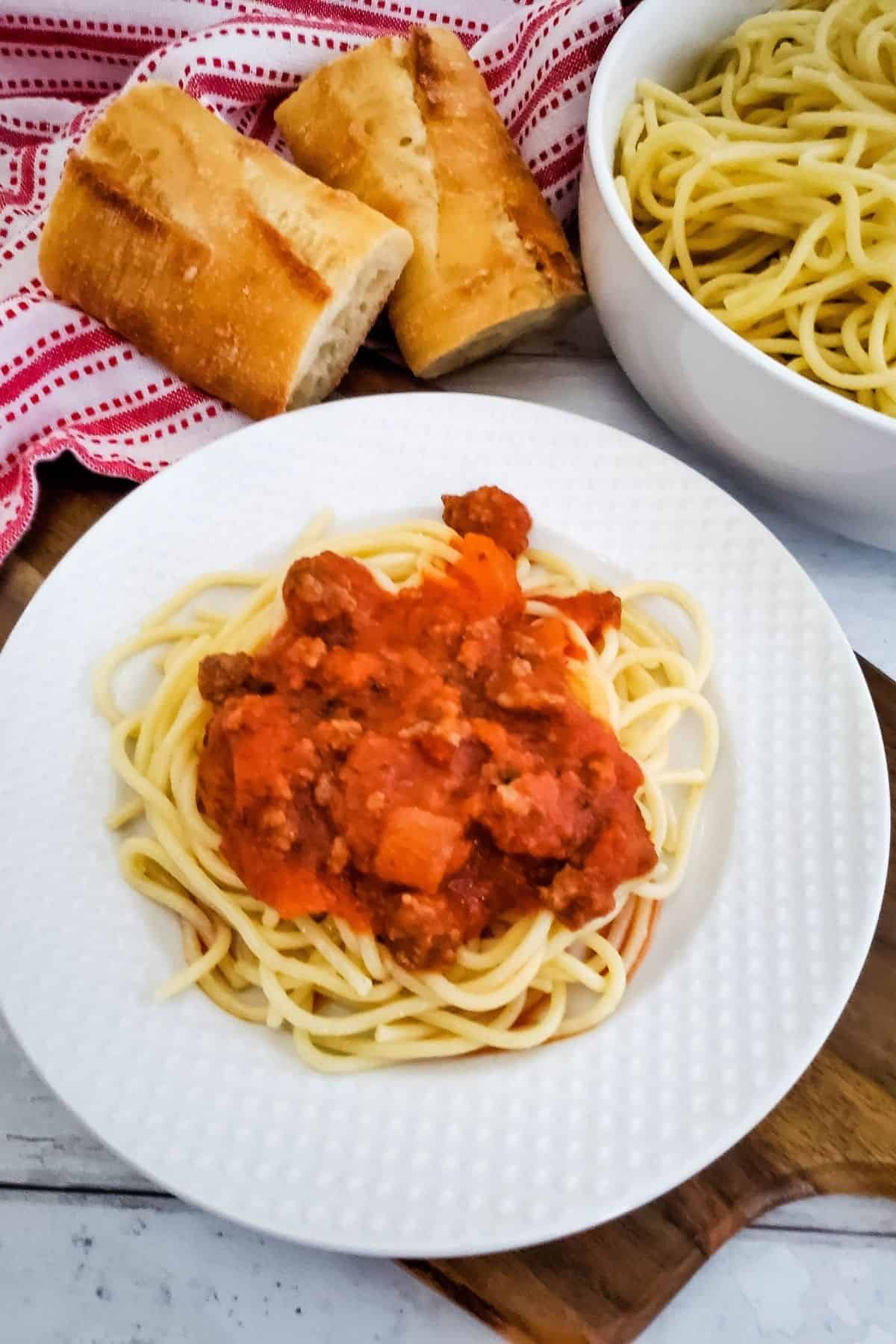 Spaghetti served on a plate with a side of bread and a bowl of pasta in the background.