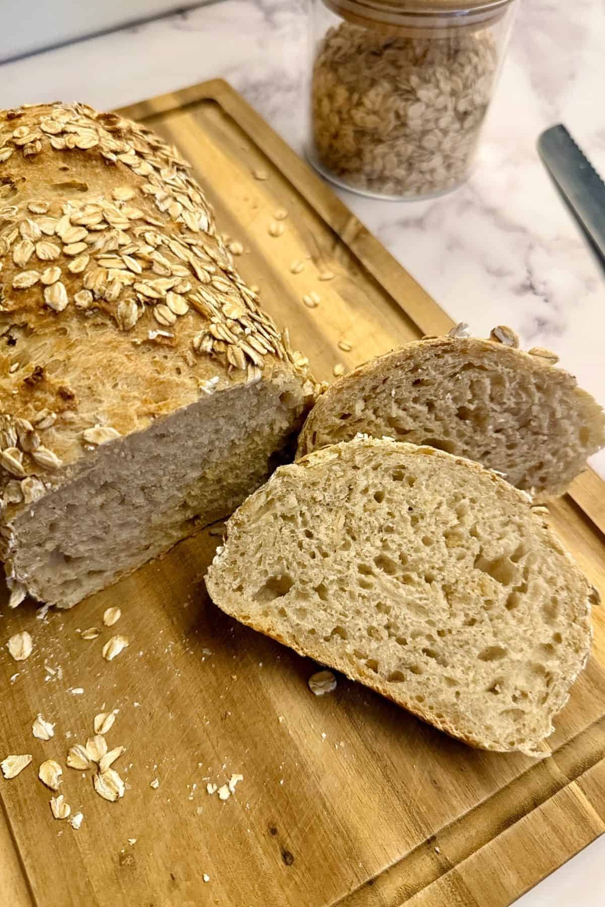Freshly baked loaf of bread with a slice beside it on a wooden board, garnished with oats.