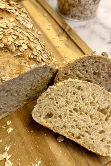 Freshly baked loaf of bread with a slice beside it on a wooden board, garnished with oats.
