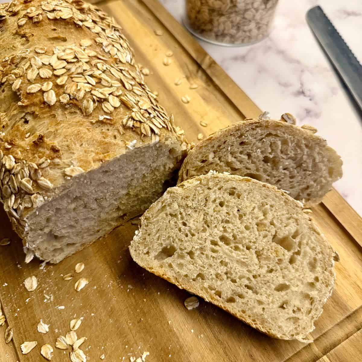 Freshly baked loaf of bread with a slice beside it on a wooden board, garnished with oats.