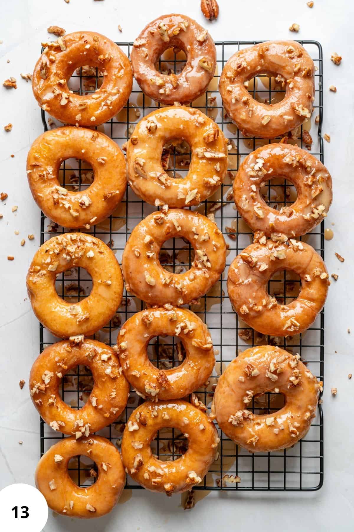 Freshly fried maple glazed donuts being topped with chopped nuts on a wire cooling rack.