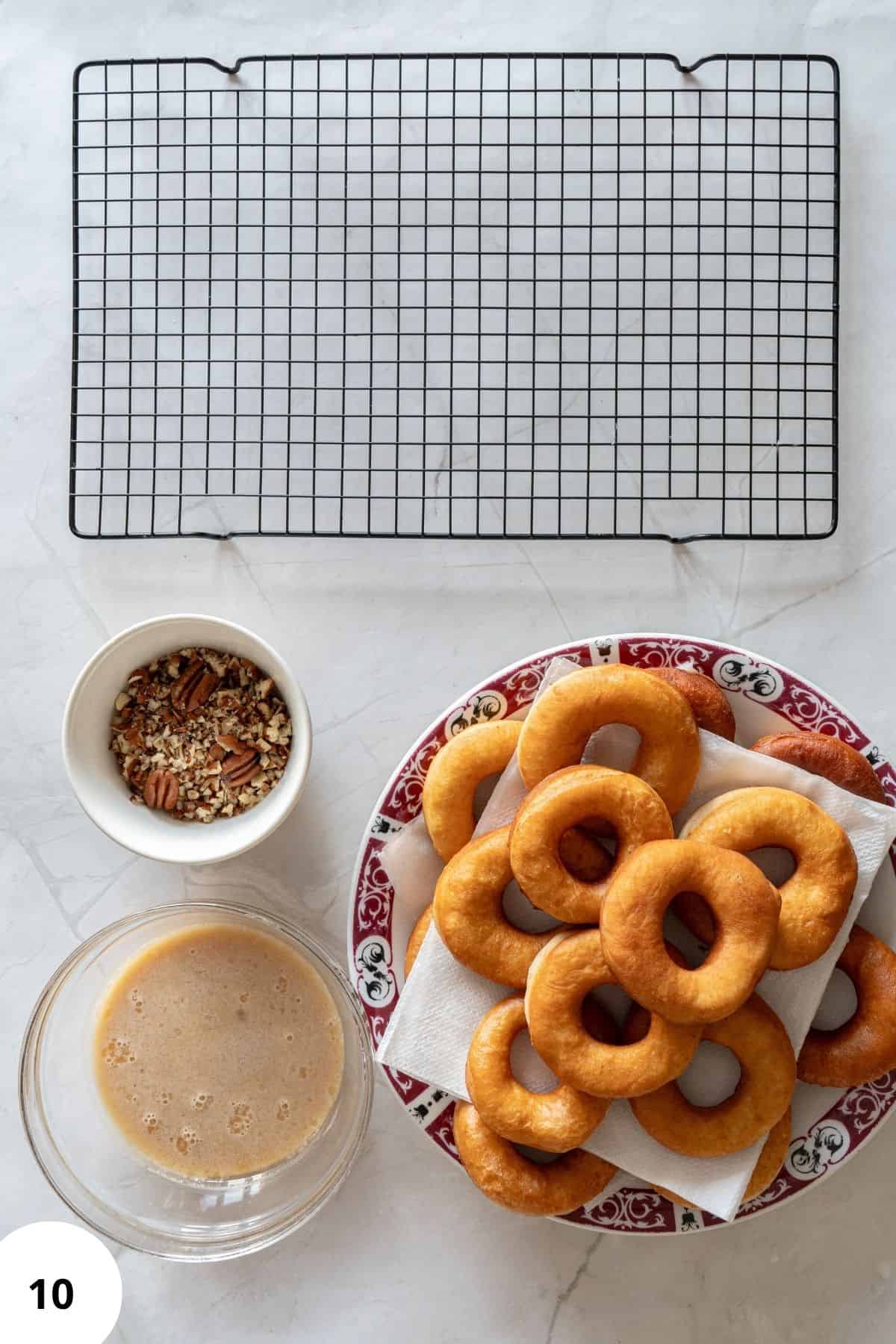 Maple glazed donuts on a plate with parchment paper being stacked beside a wire cooling rack and bowls of ingredients.