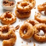 Maple glazed donuts on a surface scattered with chopped nuts.
