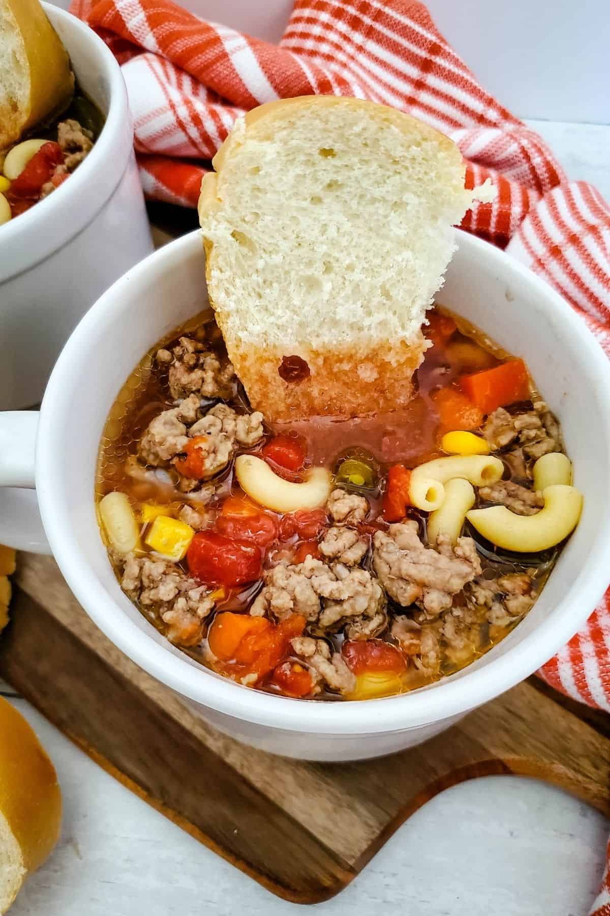Close-up photo of a bowl of cheesy cheeseburger potato soup on a rustic wooden board with a table napkin, served with bread scattered around.