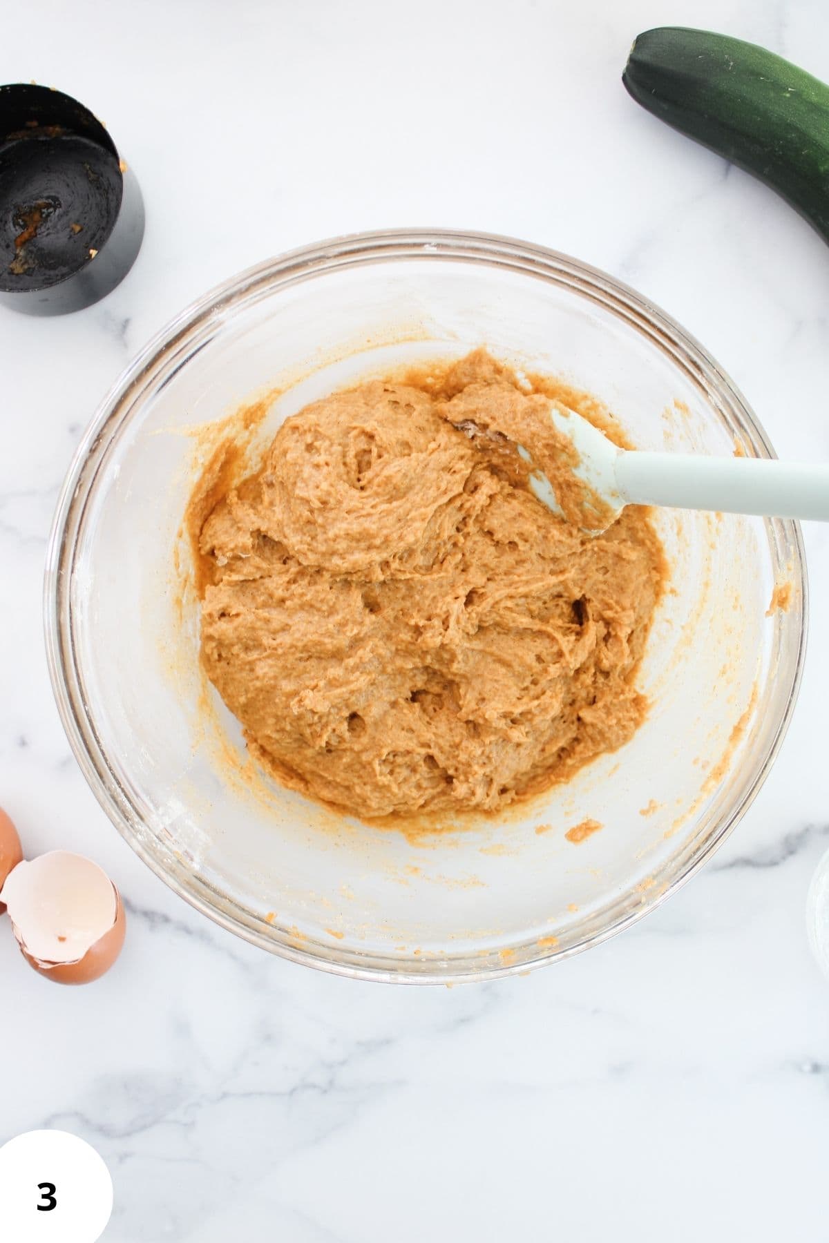 A large bowl containing zucchini muffin batter with shredded zucchini mixed in and a spatula resting on the edge.
