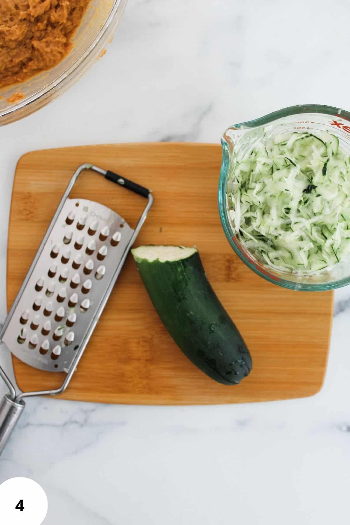 A cutting board with sliced zucchini, a grater, and a bowl filled with freshly shredded zucchini.