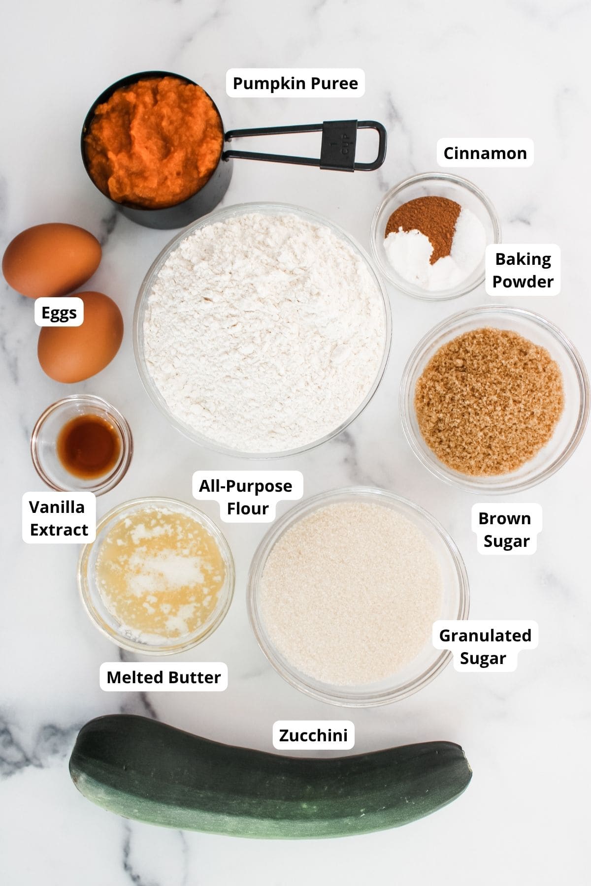 Ingredients for pumpkin zucchini muffins arranged neatly on a surface in separate bowls and containers, including flour, spices, sugars, pumpkin puree, eggs, and shredded zucchini.
