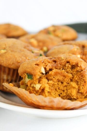 Close-up of a pumpkin zucchini muffin showing the soft, moist interior with visible zucchini and pumpkin pieces with more muffins on the background.