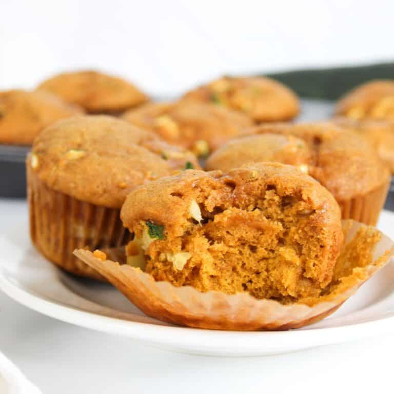 Close-up of a pumpkin zucchini muffin showing the soft, moist interior with visible zucchini and pumpkin pieces with more muffins on the background.