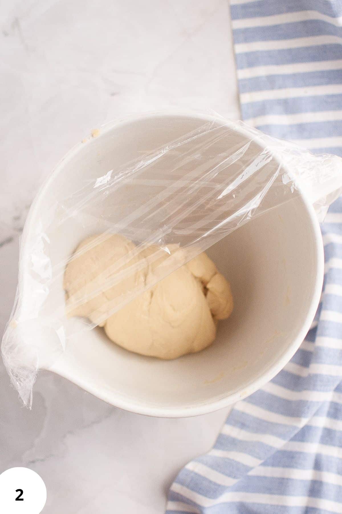 Sourdough brioche dough in a mixing bowl, half covered with plastic wrap, resting for fermentation.