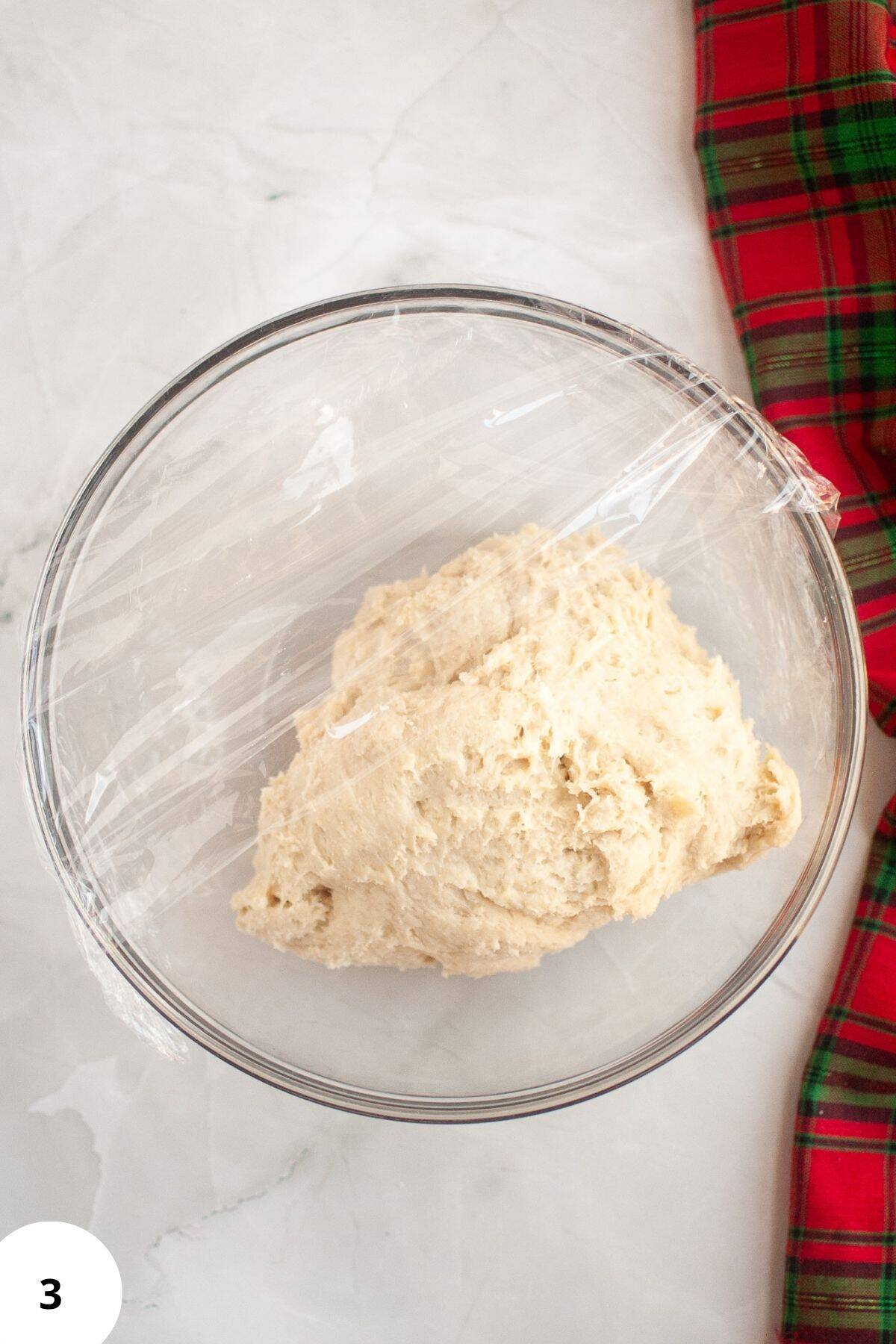 Bread dough resting in a bowl, partially covered with plastic wrap during the proofing process.