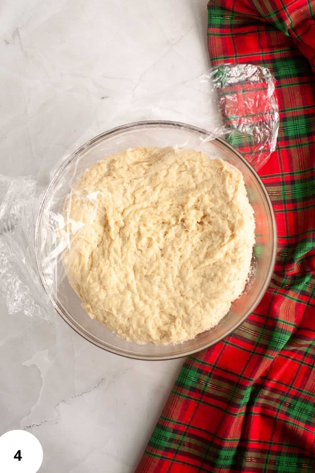 Bread dough resting in a bowl, partially covered with plastic wrap during the proofing process.