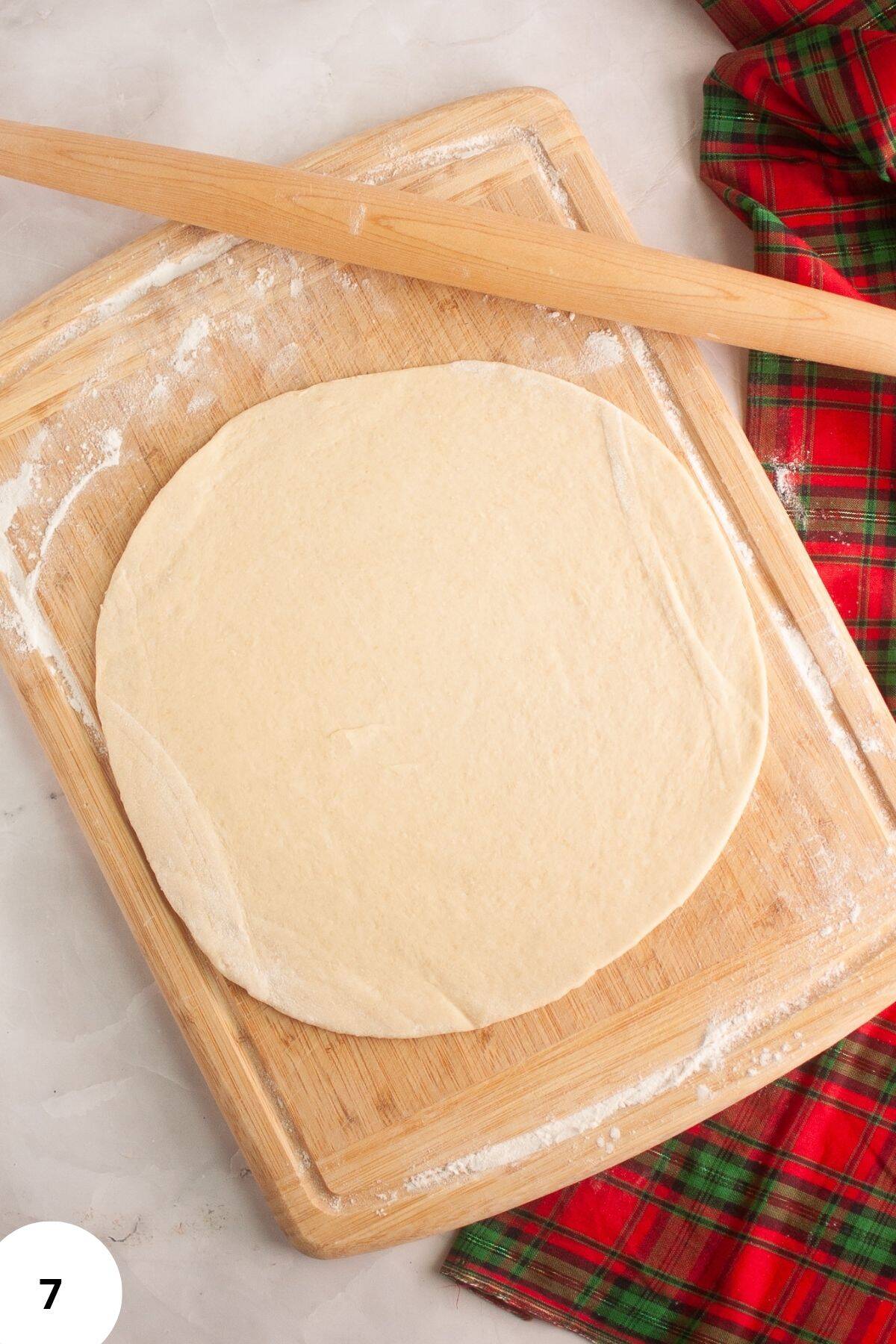 Dough being flattened on a floured wooden board with a rolling pin resting on the side.