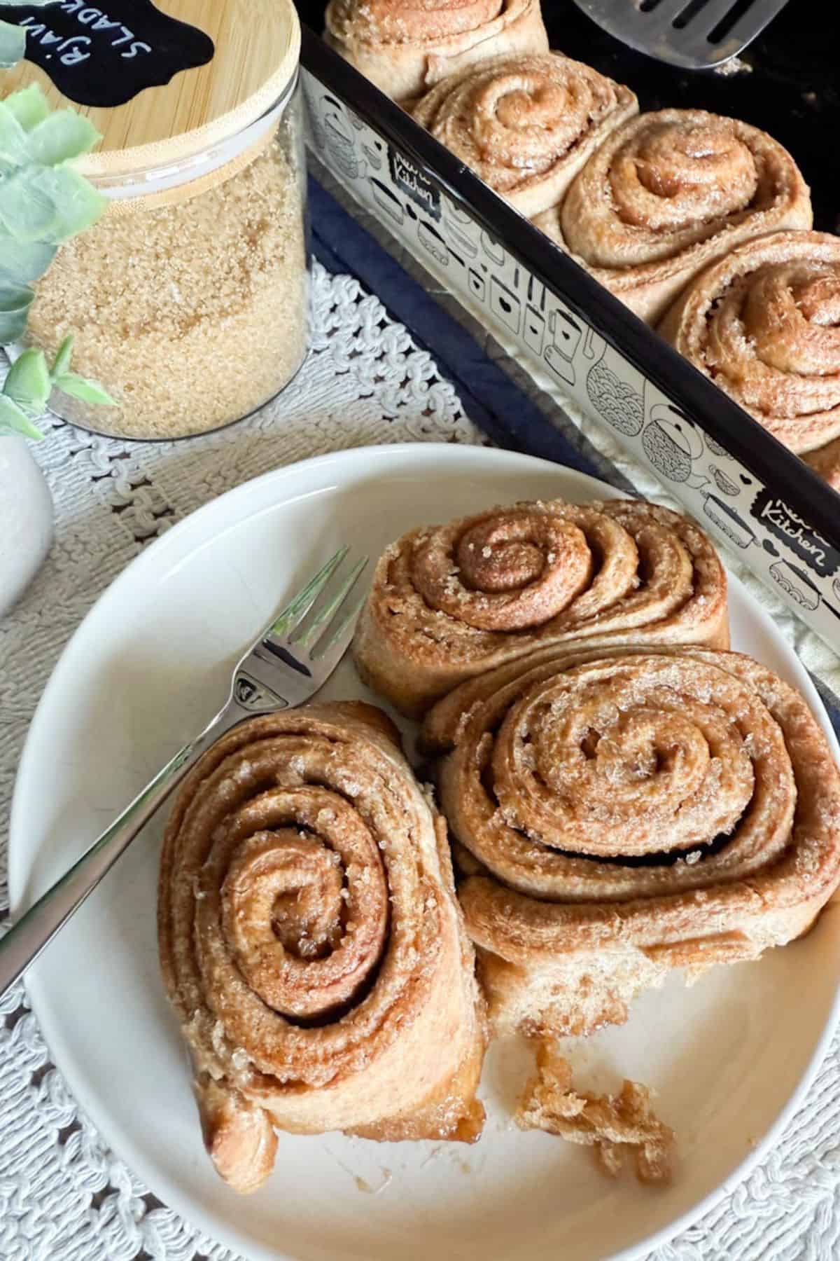 Freshly baked sourdough cinnamon rolls served on a plate beside a baking pan full of rolls.