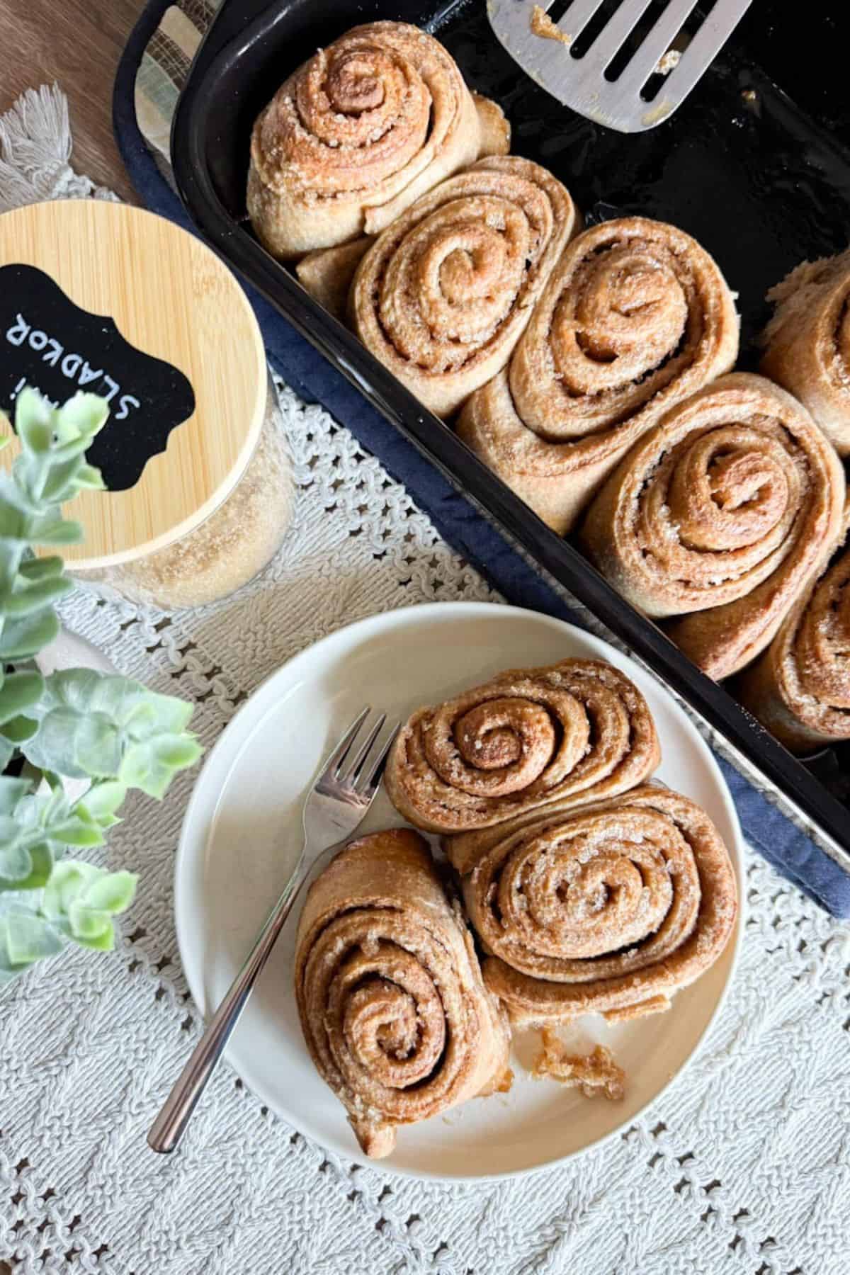 Freshly baked sourdough cinnamon rolls served on a plate beside a baking pan full of rolls.