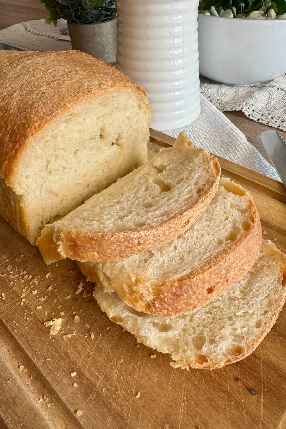 A loaf of bread with slices cut, displaying a soft, airy interior, on a wooden cutting board with kitchen decor in the background.