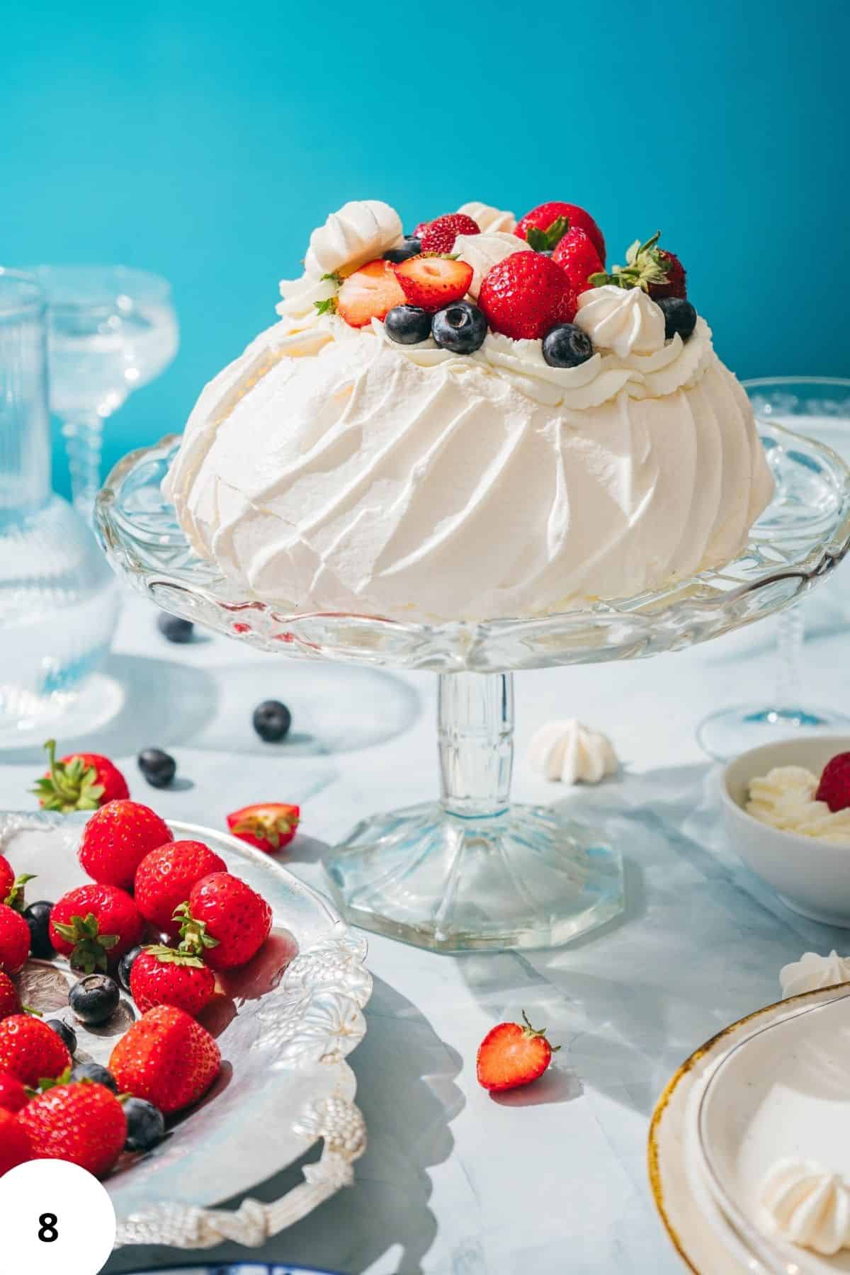 Bright and colorful pavlova topped with fresh berries and whipped cream on a glass cake stand.