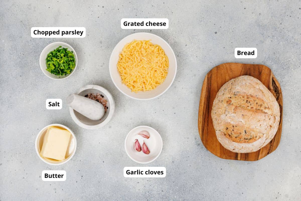 Fresh bread on a wooden board with chopped parsley, shredded cheese, and garlic cloves.