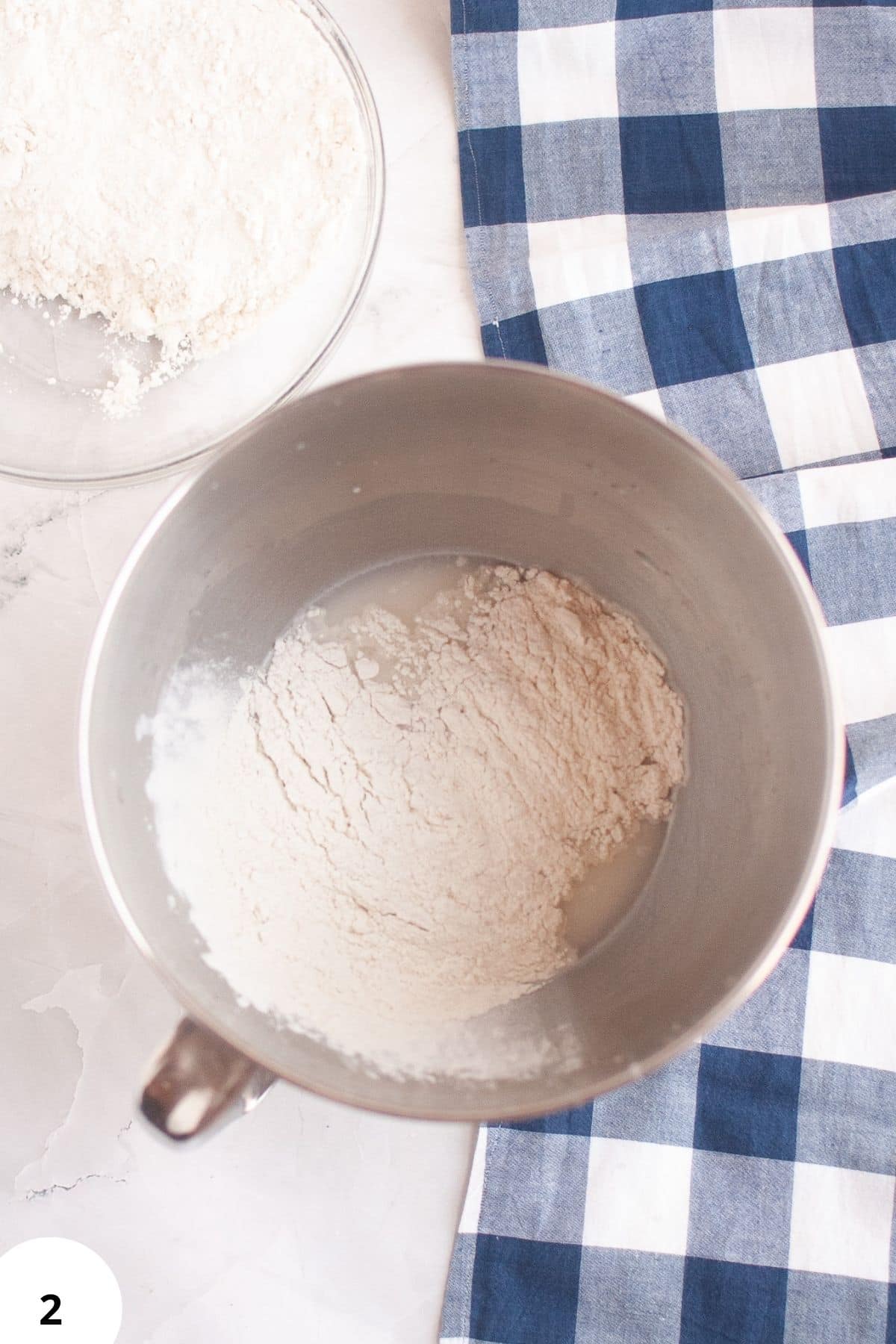 Alt text: Dough rising in a metal bowl with flour, blue checkered cloth nearby.