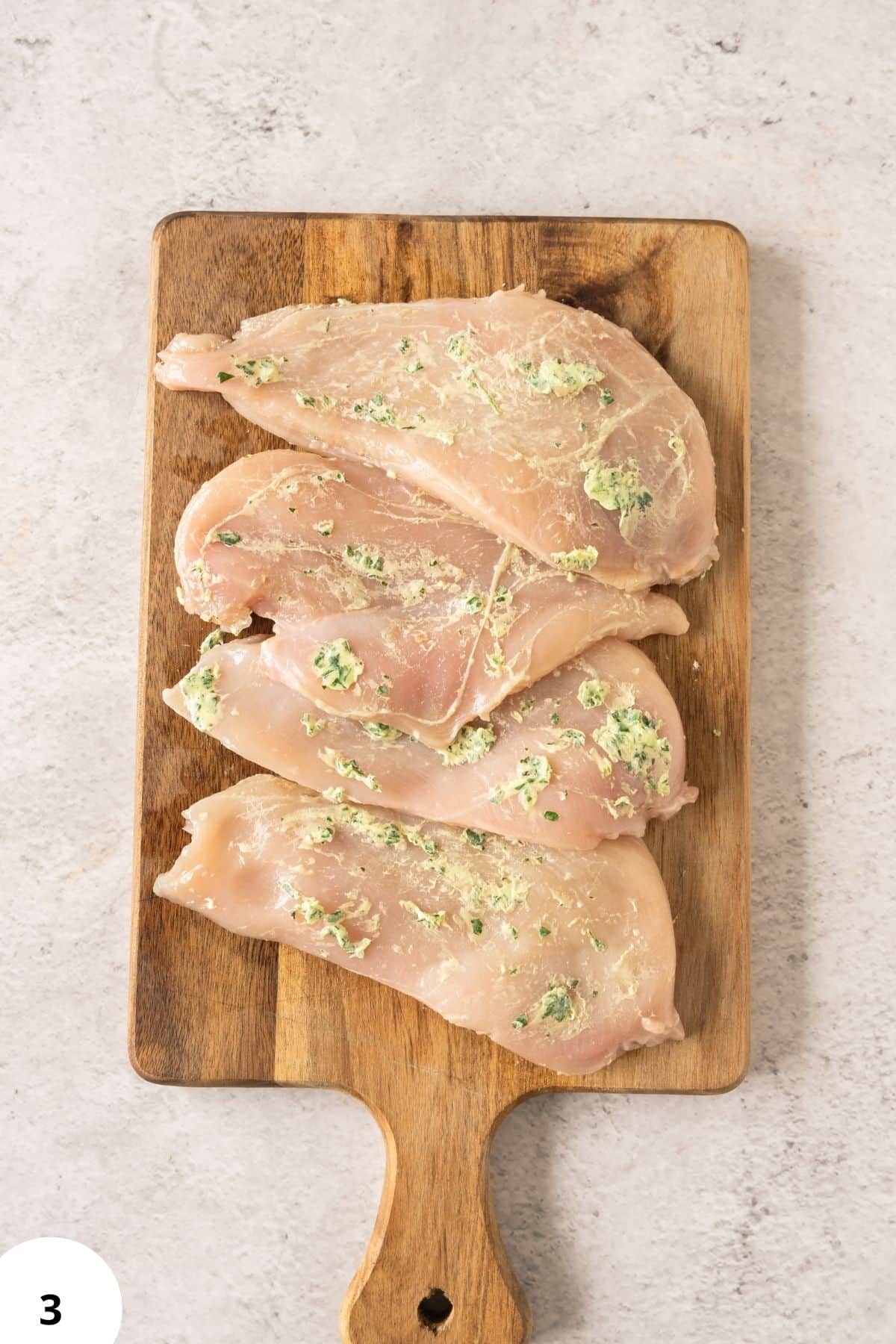 Fresh chicken breast fillets with garlic and herb coating on wooden cutting board.