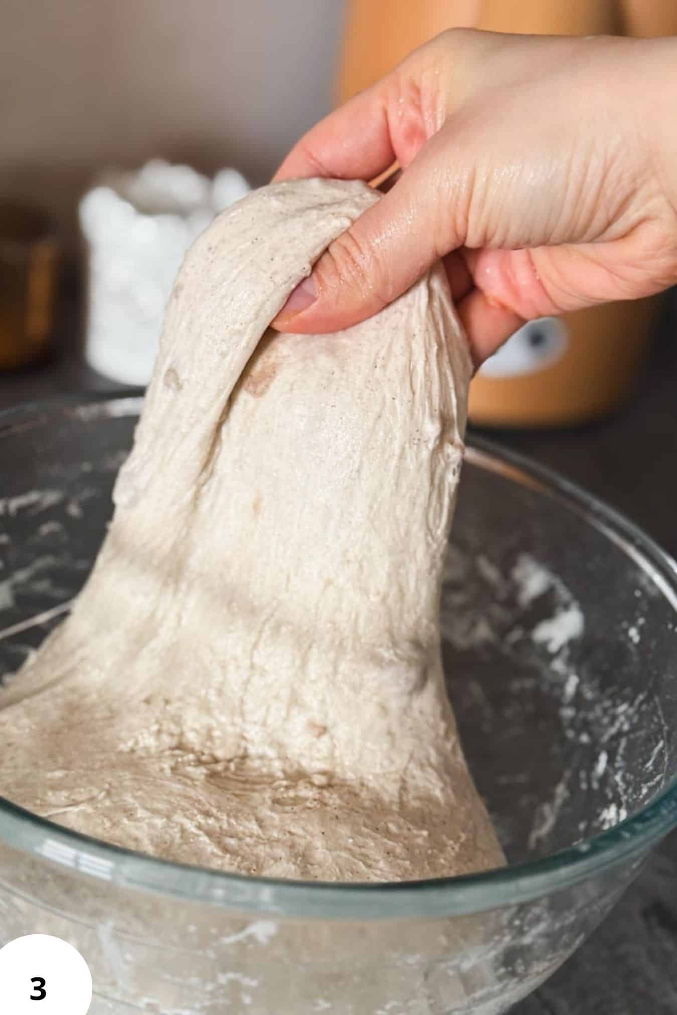 Hand stretching buckwheat sourdough dough in a glass bowl for bread fermentation.