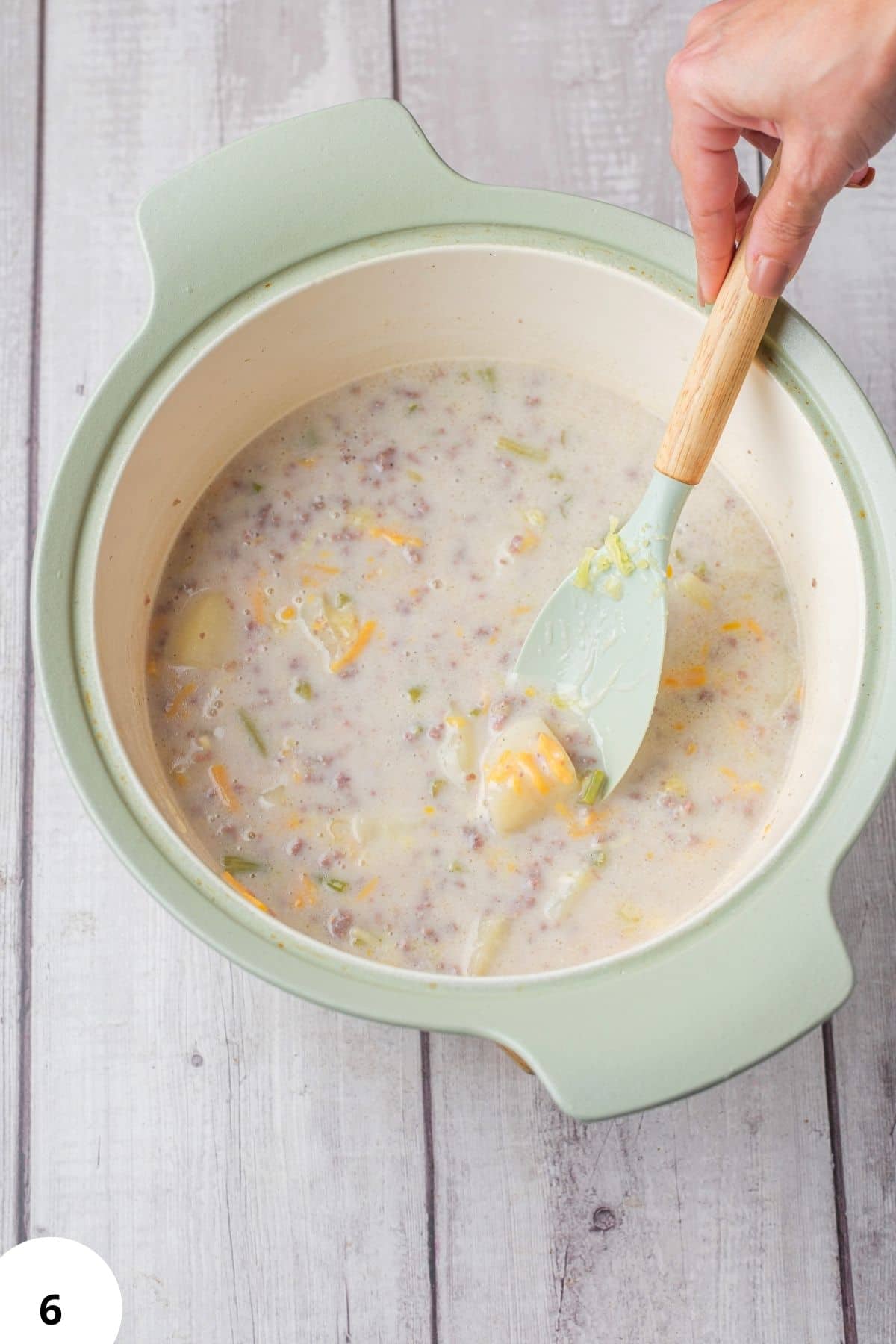 Hearty hamburger and potato soup in a light green pot with a hand stirring.