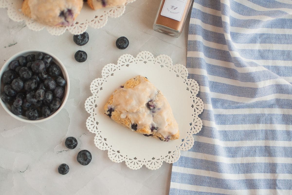 Blueberry cream scone on a decorative doily with fresh blueberries and a striped cloth nearby.