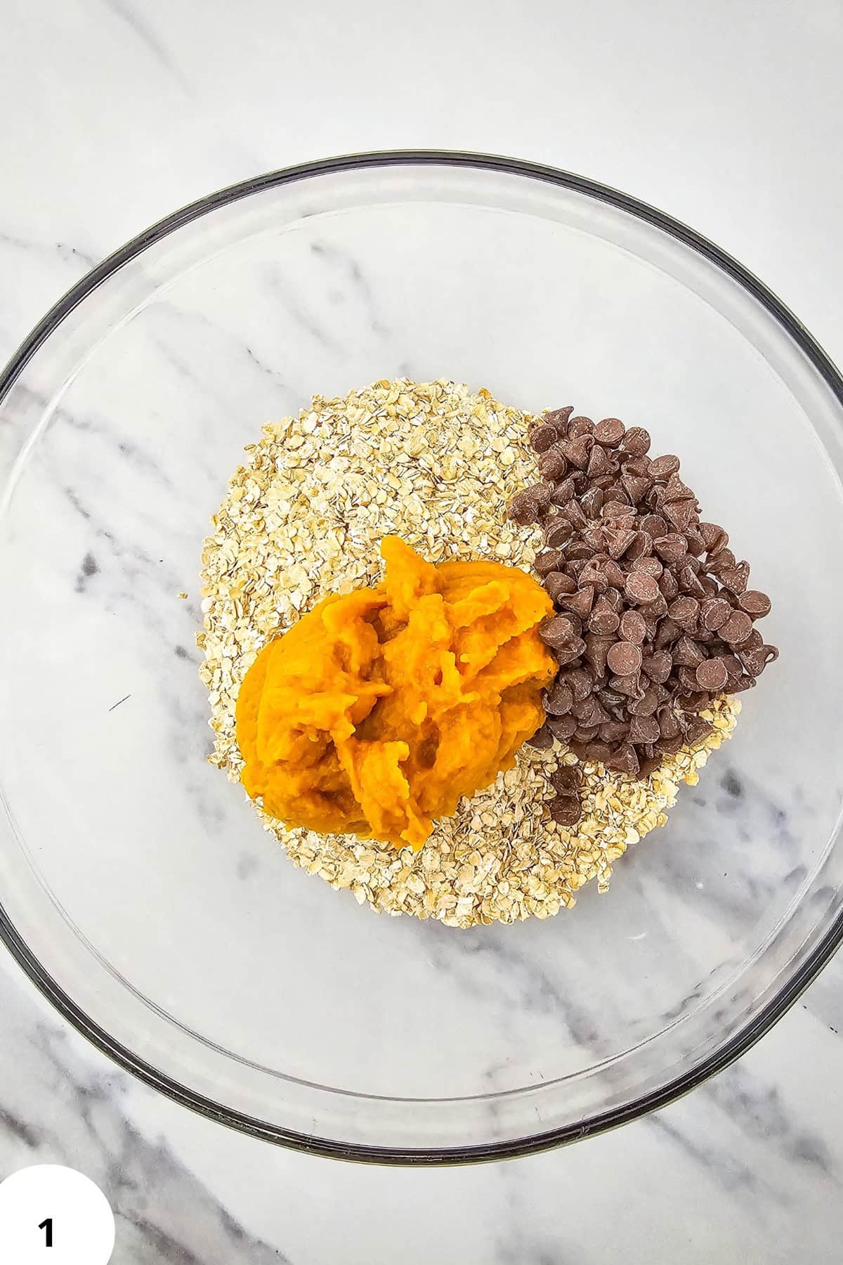 Ingredients for pumpkin oatmeal cookies in a glass bowl, including oats, chocolate chips, and pumpki.