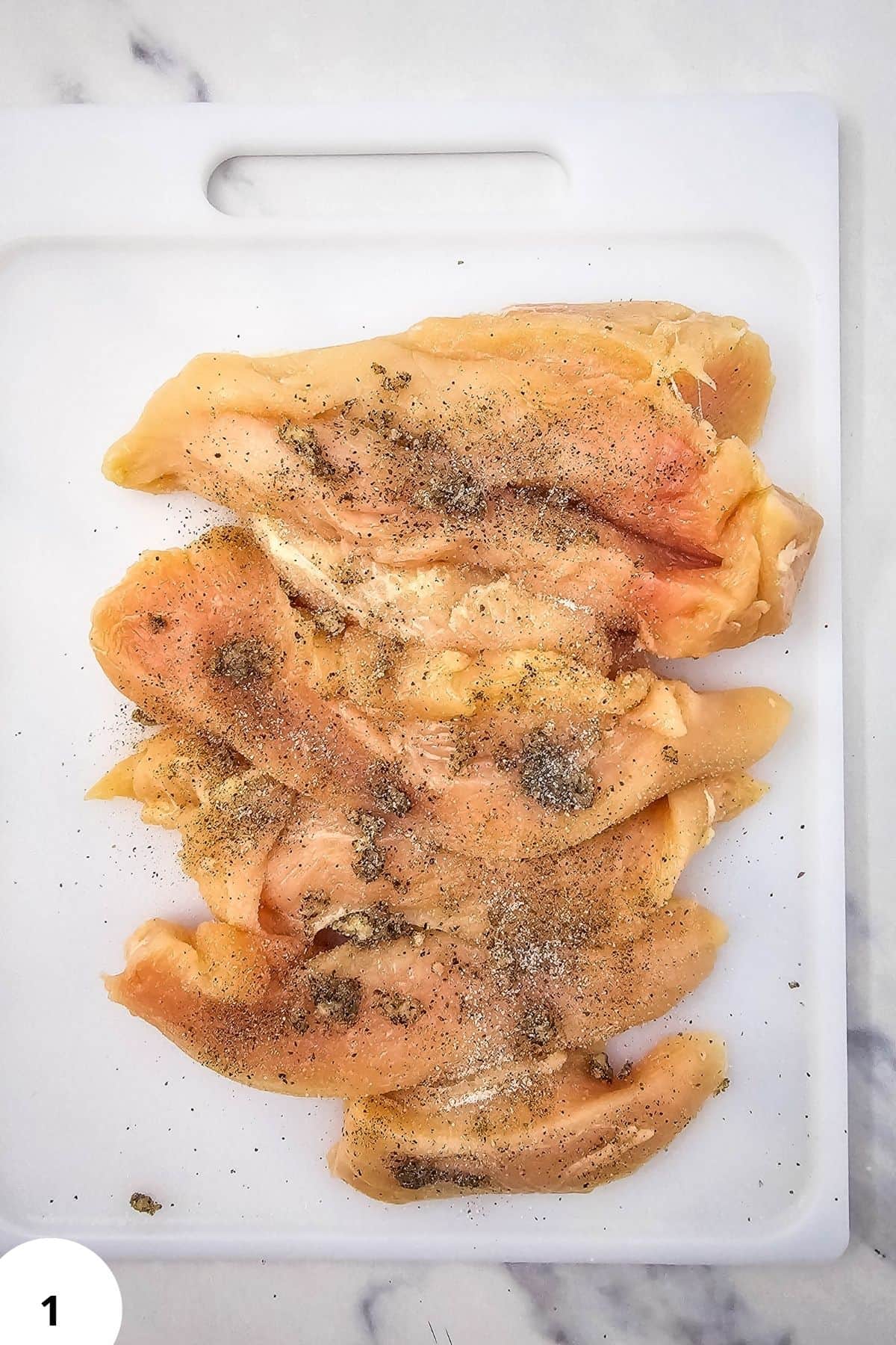 Raw chicken breasts seasoned with black pepper on a white cutting board.