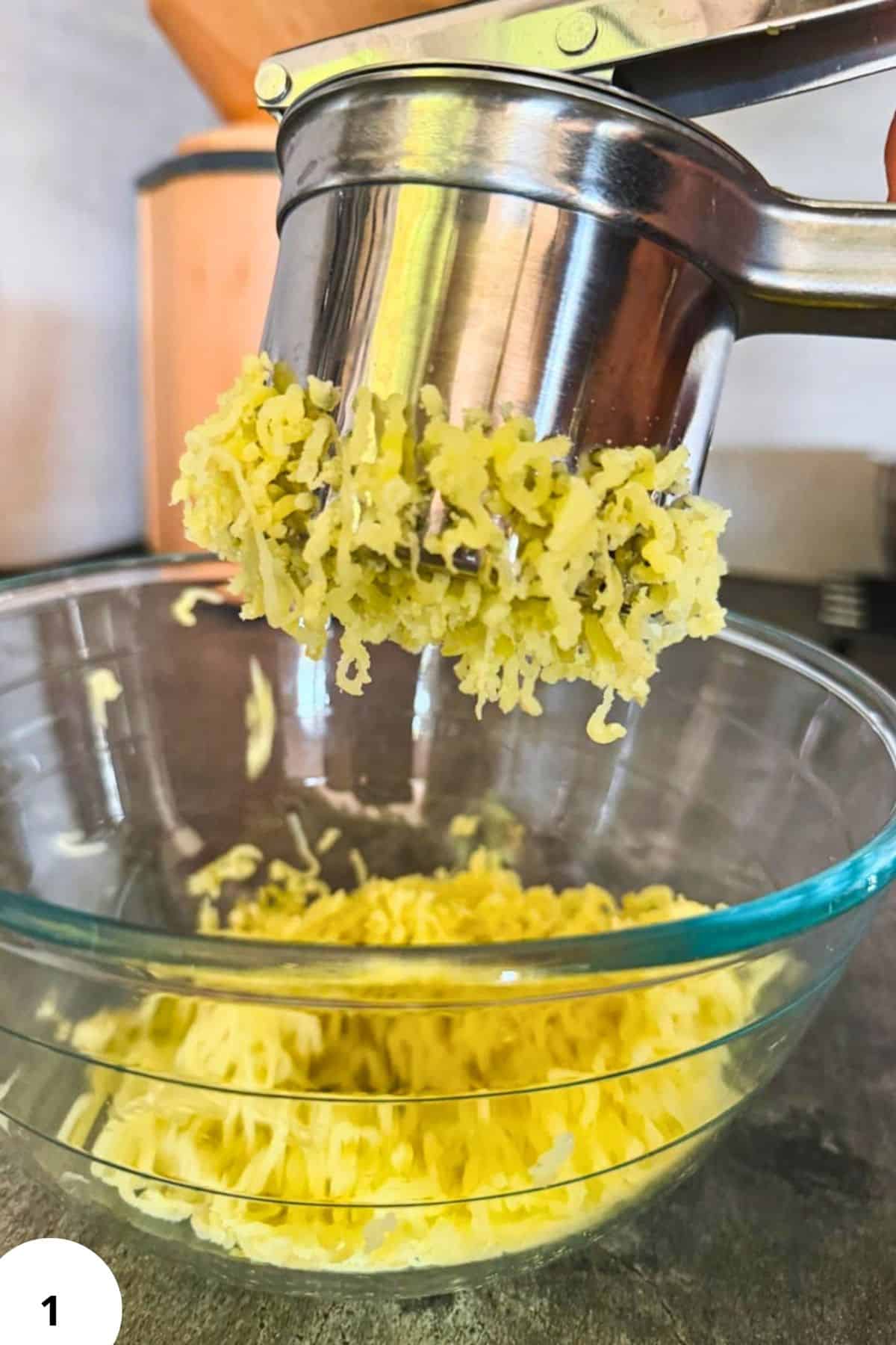Hand-cranked potato ricer pressing freshly milled potatoes into a glass bowl.