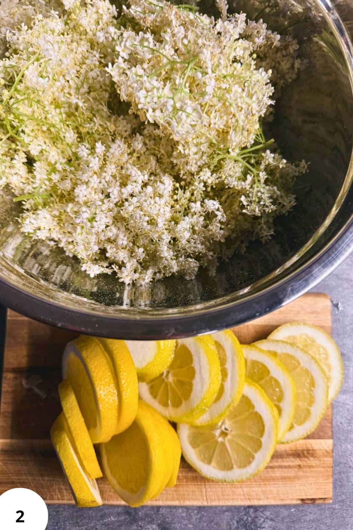 Elderflowers in a bowl with lemon slices on a wooden cutting board, ready for culinary use.