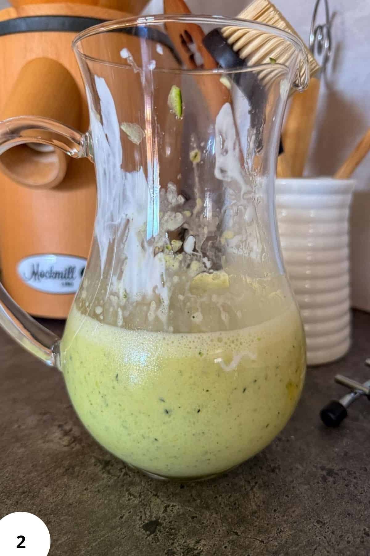 Freshly blended green smoothie in a glass pitcher with kitchen utensils in background.