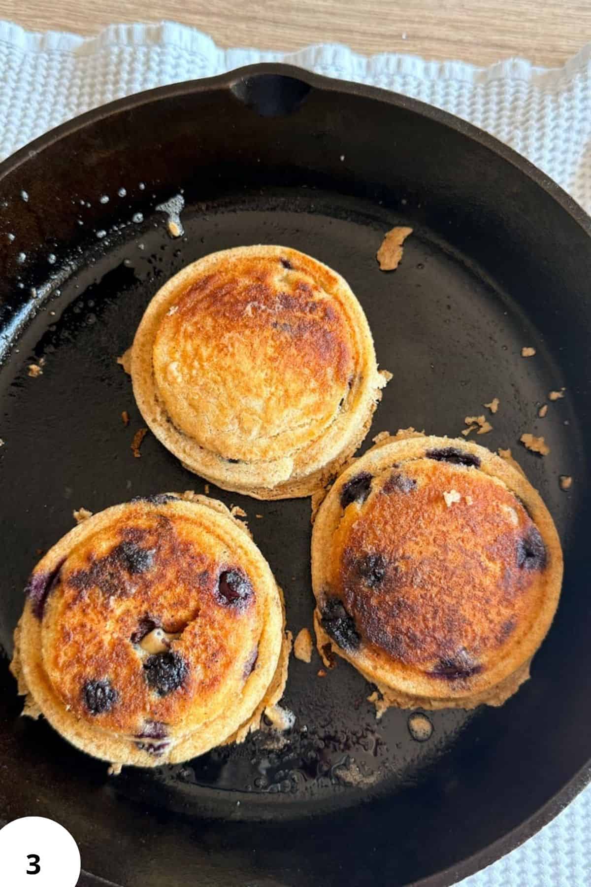 Three golden sourdough pancakes with blueberries in a black skillet.