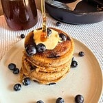 Stack of blueberry pancakes topped with butter and syrup, surrounded by blueberries.