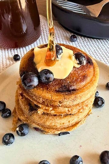Stack of blueberry pancakes topped with butter and syrup, surrounded by blueberries.