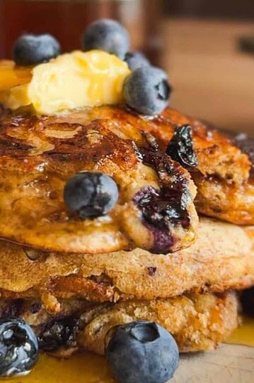 Stack of blueberry pancakes topped with butter, blueberries, and syrup.