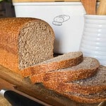 Sliced homemade bread on a wooden cutting board with a bread box in the background.