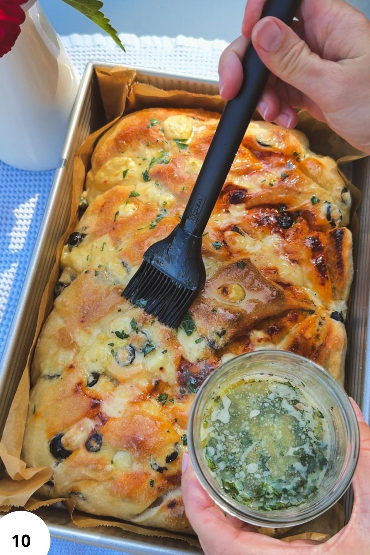 Freshly baked sourdough focaccia with tomatoes, olives, and pesto, being brushed with olive oil.