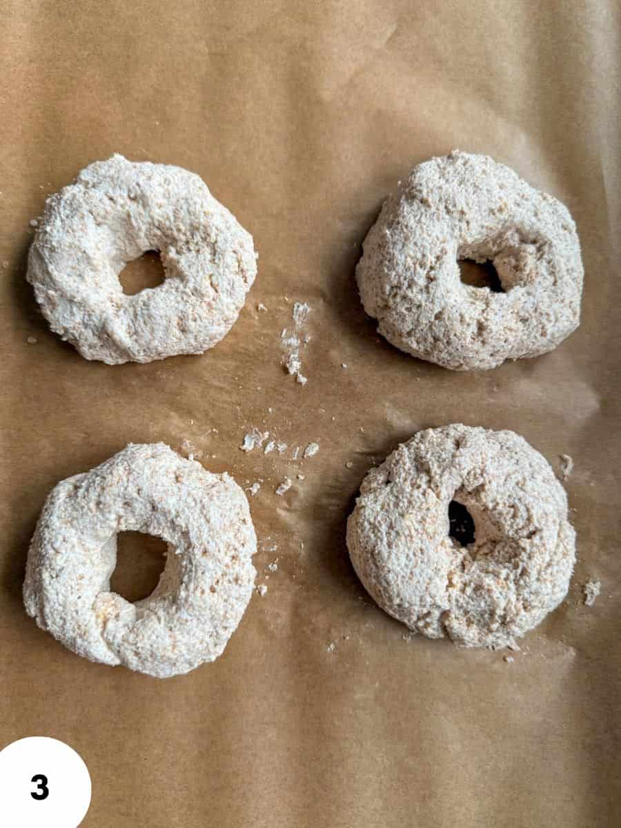Four bagels coated with flour on parchment paper for baking.