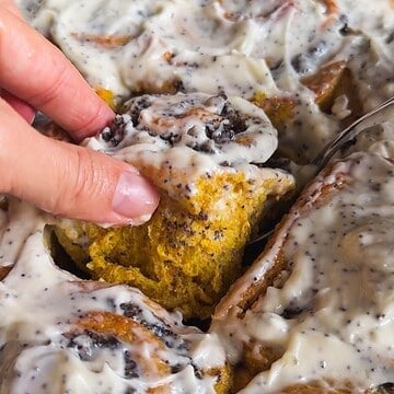 Close-up of a cinnamon roll cake with creamy icing and a hand holding a piece.