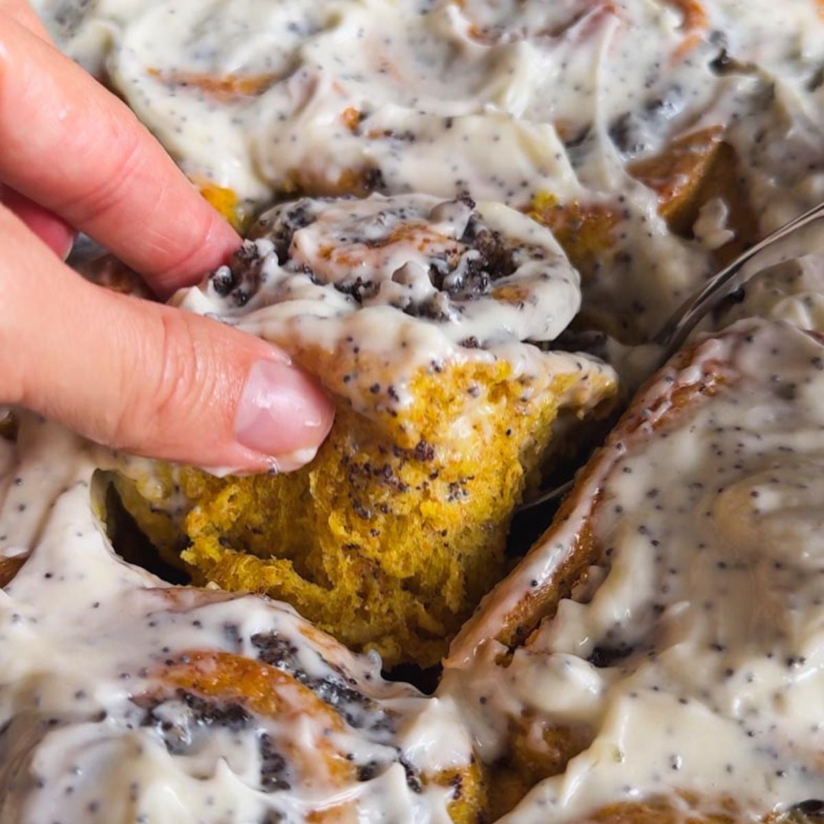 Close-up of a cinnamon roll cake with creamy icing and a hand holding a piece.
