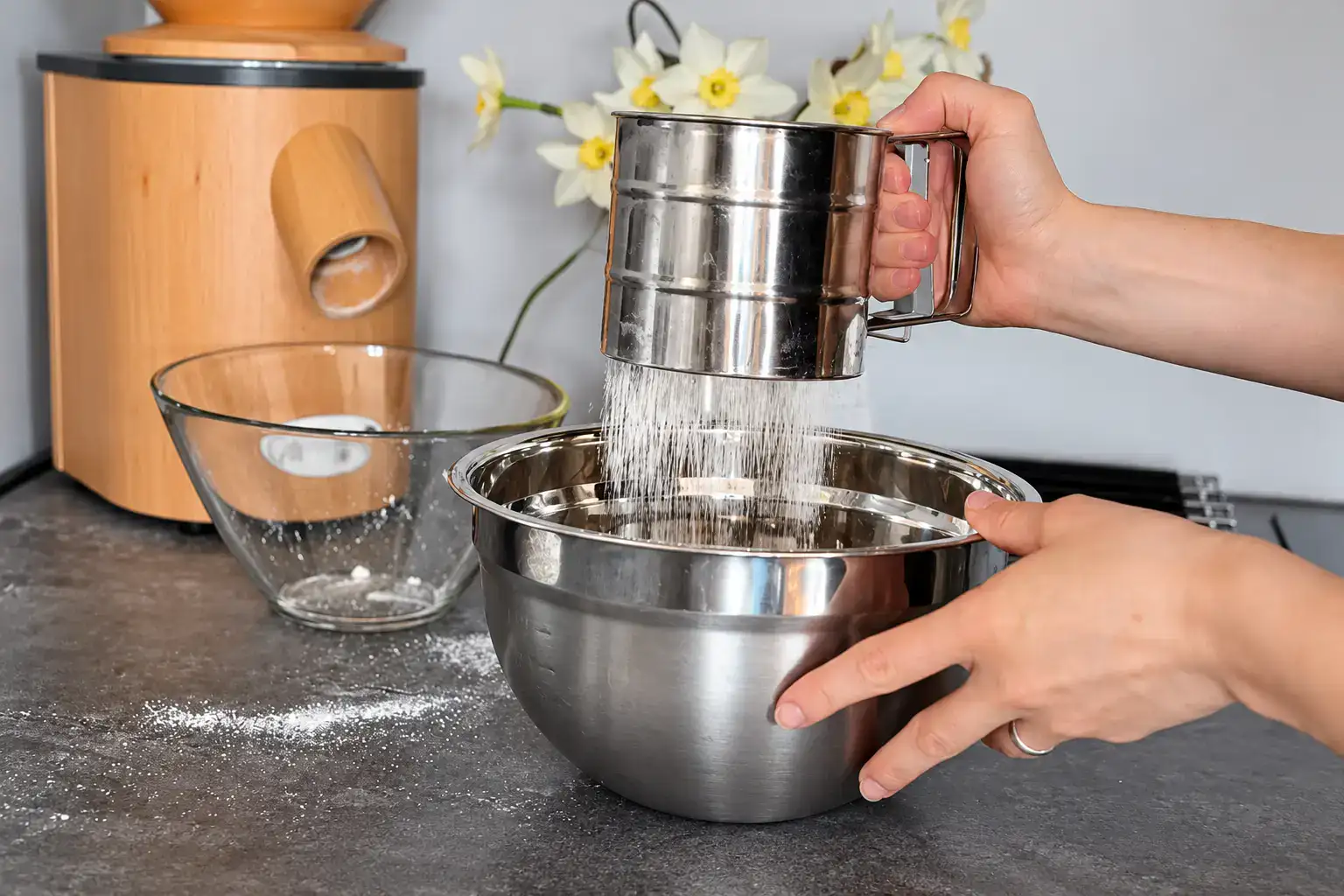 Freshly milled flour being sifted through a metal flour sifter into a stainless steel mixing bowl