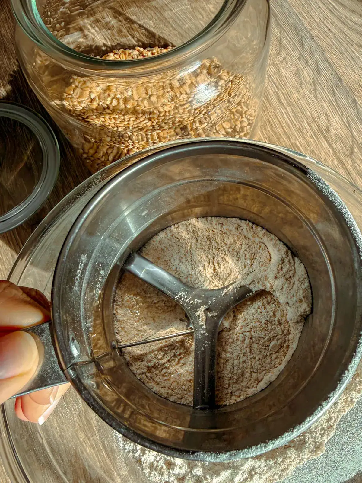 Hand sifter filled with fresh milled flour held over a glass bowl, wheat berries in a jar behind it